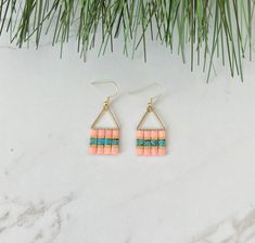 the pink and green earrings are hanging from gold hooks on a white marble countertop