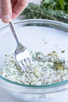 a person holding a fork in a glass bowl filled with white sauce and herbs on the side