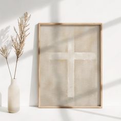 a white vase with some flowers in it next to a framed cross on the wall