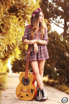 a woman in a dress holding a guitar on a sidewalk with trees and bushes behind her