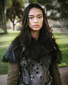 a woman with long black hair wearing a leather jacket and standing in front of trees