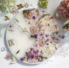 a clock with flowers on it sitting next to some pink and white flowers