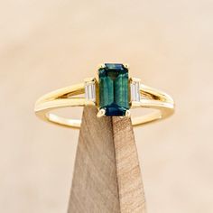 an emerald and diamond ring sitting on top of a wooden block in front of a beige background