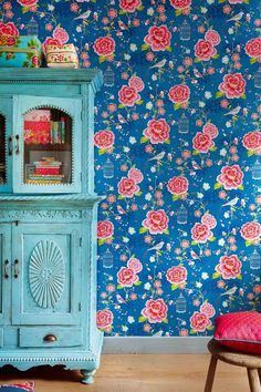 a blue cabinet sitting in front of a wall with flowers on it and a pink chair next to it