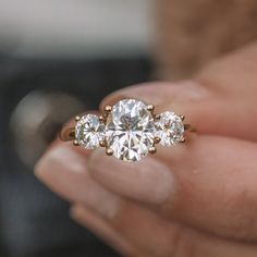 a woman's hand holding an engagement ring with three stones on it and the center stone is surrounded by smaller round diamonds