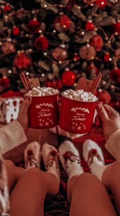 two people holding red mugs filled with marshmallows in front of a christmas tree