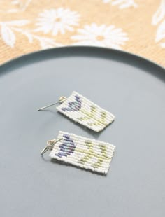 two white and blue beaded earrings sitting on top of a black plate next to a flowered tablecloth