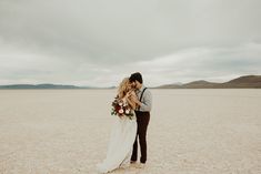 a bride and groom kissing in the middle of an empty desert field with mountains in the background