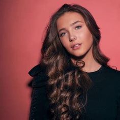 a woman with long hair standing in front of a red wall wearing a black top