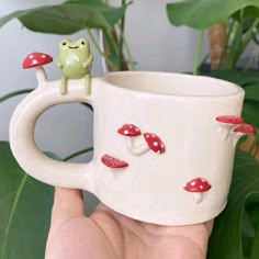 a hand holding a white coffee cup with red mushrooms on it and a frog figurine sitting on top