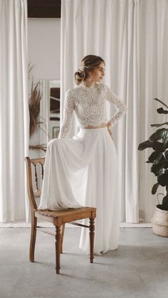 a woman standing in front of a window wearing a long sleeved wedding dress with an open back