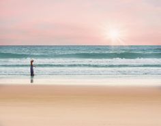 a woman standing on top of a beach next to the ocean under a pink sky