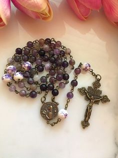 "Handmade bronze St. Teresa of Avila rosary made with 6mm purple amethyst gemstone beads and 8mm purple & white floral ceramic beads. The vintage inspired bronze Saint Teresa center measures 13/16\". The crucifix measures approximately 1 3/4\"." Lavender Spiritual Jewelry With 8mm Beads, Spiritual Lavender Jewelry With 8mm Beads, Lavender Spiritual Jewelry With Beads, Purple Rosary With 8mm Beads, Purple Rosary With 8mm Round Beads, Handmade Spiritual Purple Rosary Bracelet, Handmade Adjustable Purple Rosary, Purple Beaded Rosary As Gift, Beautiful Rosaries