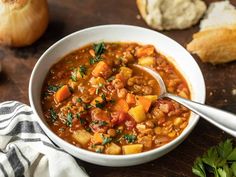 a white bowl filled with vegetable soup on top of a wooden table next to bread