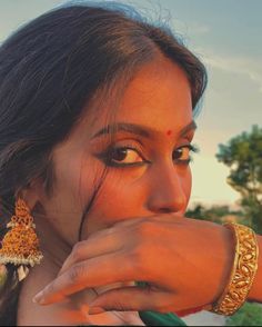 a woman wearing gold jewelry and holding her hand to her mouth while looking off into the distance