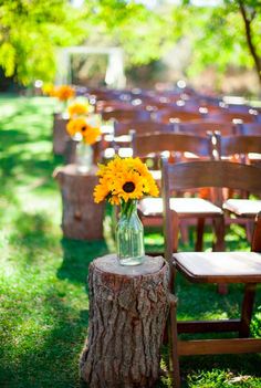 sunflowers are in a vase sitting on a tree stump at an outdoor ceremony