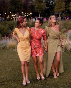 three women in dresses are standing together on the grass and smiling at each other while holding hands