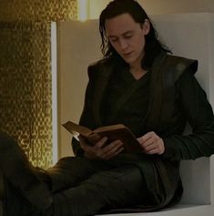 a person sitting down reading a book and looking at the camera while wearing black clothing