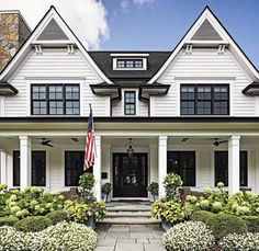 a large white house with black windows and two flags on the front porch, surrounded by landscaping