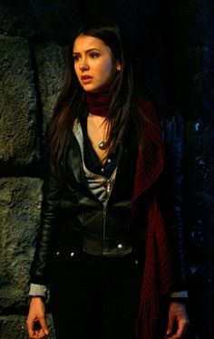 a woman standing in front of a stone wall wearing a black leather jacket and scarf