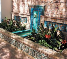 an outdoor fountain with plants and flowers in the center, next to a brick wall