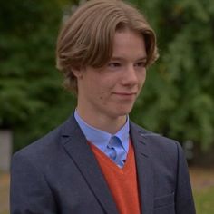 a young man wearing an orange shirt and blue blazer standing in front of trees