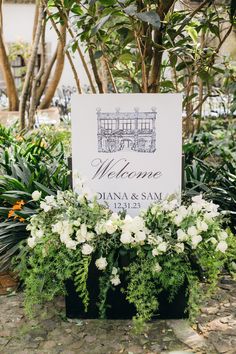 a welcome sign sitting on top of a planter filled with white flowers and greenery