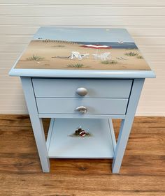 a small blue table with a painting on the top and bottom drawer, sitting on a hard wood floor