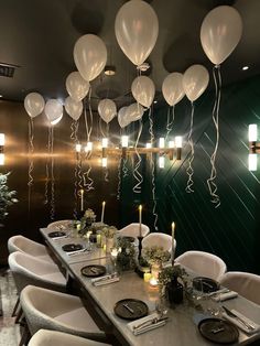 a long table with white balloons hanging from the ceiling and place settings for dinner guests