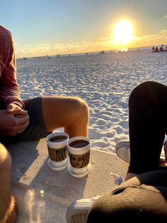 two people sitting on the beach with cups of coffee