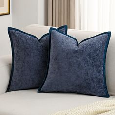 two blue velvet pillows sitting on top of a white couch next to a beige blanket