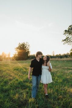 a man and woman are standing in the grass with their arms around each other as the sun sets behind them