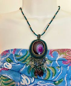 When I ran across this cabochon I had to have it. I crafted this necklace in bead embroidery with deep teal beads to compliment the intense purple color of the cabochon. I added sweet pressed glass leaves on the fringe. This is a one of a kind piece and if you are a purple lover, like me, it will not disappoint! The pendant from loops to fringe measures 4 inches long and 2 inches wide. The seed bead necklace is 21 inches long and features a gold tone lobster claw clasp. Bohemian Oval Beaded Necklaces, Bohemian Oval Beaded Necklace, Handmade Purple Pendant Beaded Necklace, Handmade Purple Beaded Pendant Necklace, Handmade Oval Beaded Bohemian Necklaces, Handmade Bohemian Oval Beaded Necklaces, Bohemian Beaded Necklaces With Cabochon For Gifts, Oval Beaded Gems For Jewelry Making, Embroidery Necklace