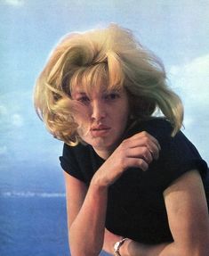 a woman with blonde hair is posing for a photo near the ocean and has her hand on her shoulder