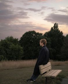 a man sitting on top of a wooden bench next to a field with trees in the background