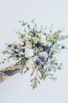 a person holding a bouquet of white and blue flowers in their hand with greenery on the side