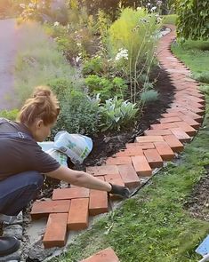 a woman kneeling down to pick up bricks from the ground in front of her garden