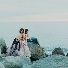 two women sitting on rocks near the ocean with their backs to each other and one holding a baby