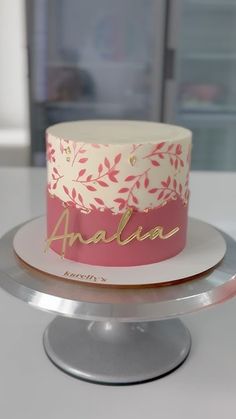 a pink and white cake with gold lettering on it sitting on a silver plate in front of a window