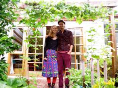 a man and woman standing next to each other in front of a house surrounded by plants