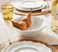 two pears in a white bowl on a table with plates and wine bottles behind it