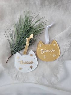 two christmas ornaments are laying on a white sheet and one has a pine branch in the corner