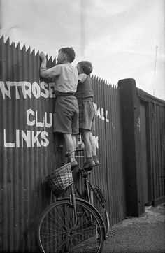 black and white photograph of two boys on top of a bike leaning against a fence