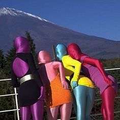 three mannequins dressed in different colored outfits standing next to a fence with a mountain in the background