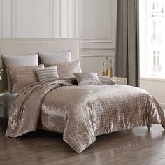 a bed with gold comforter and pillows in a white room next to a window