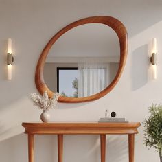 a round mirror on the wall above a wooden table with vase and flowers in it