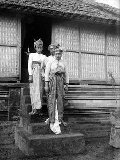 two women standing on steps in front of a building with bamboo screens behind them and one woman wearing a headdress