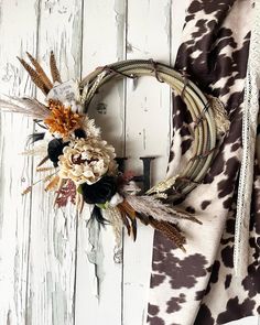 a wreath with dried flowers and feathers hanging on a white wooden wall next to a cow print scarf