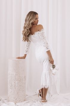 the pregnant woman is posing in her wedding dress and holding a flower bouquet while leaning against a white backdrop