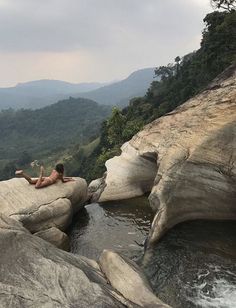 a woman laying on top of a large rock next to a river in the mountains